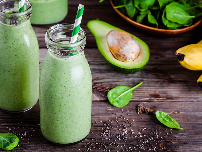 Zwei Trinkgläser mit grünen Smoothie und grün-weißen Strohhalmen. Avokado, Spinat und Banane im Hintergrund. © shutterstock Inc. 