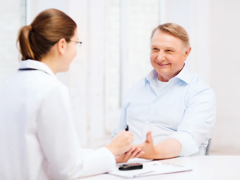 Älterer Herr in blauen Hemd sitzt gegenüber einer jungen Ärztin, die ihm Blutzucker misst. © Shutterstock Inc.